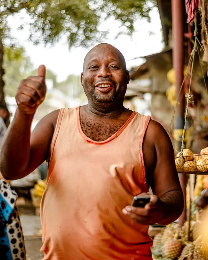 A shopkeeper gives a thumbs up at his stall, phone in hand.