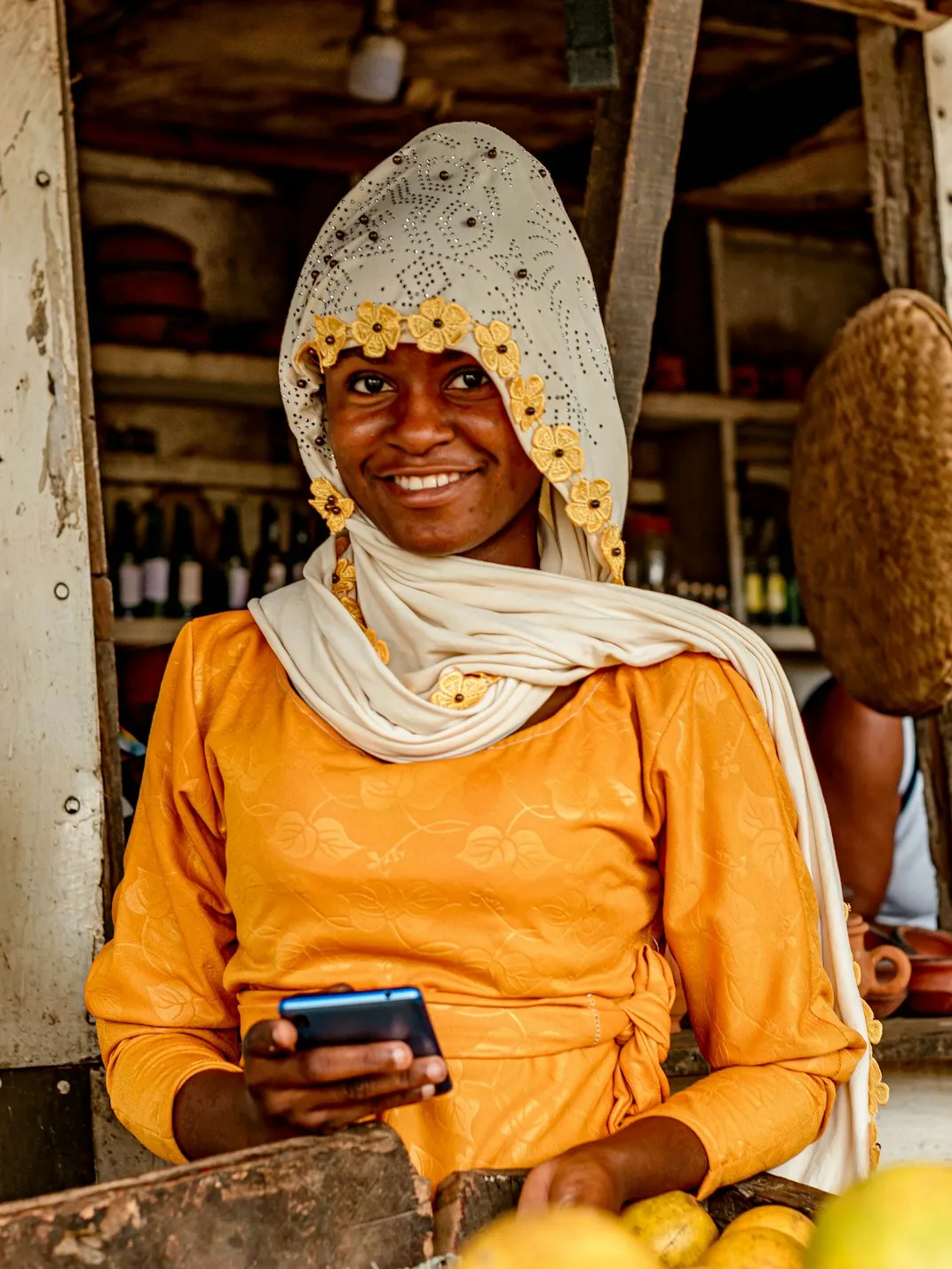 A trader in a yellow dress smiles at her stall, a phone in her hand.